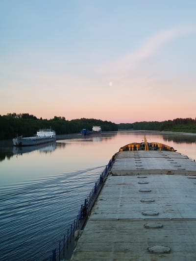 This serene image captures a twilight view from the deck of a large cargo vessel navigating a calm river, likely near Volokov Most, Russia. The scene unfolds during either sunrise or sunset, evidenced by the soft, pastel hues of pink, peach, and light blue in the sky, and the visible full moon high above.
In the foreground, the sturdy, flat deck of the viewer's vessel is prominent, featuring rows of circular hatches or covers. A blue railing lines the port side of the ship, and a clear, ripple-filled wake trails behind, indicating the vessel's movement through the water.
The river itself is tranquil and reflective, mirroring the soft colors of the sky. Both banks are densely lined with lush green trees, suggesting a natural and forested landscape. Further along the river, two other cargo ships are visible. On the left, a long, grey and white barge, possibly a dry cargo ship, is moving or moored, with a central cabin structure. The Cyrillic text "ПОКРОВСК" is clearly discernible on its side. Beyond it, further upstream and to the right, a second, darker blue-hulled vessel can be seen.
The atmosphere is peaceful and quiet, characteristic of an early morning or late evening journey on the water.