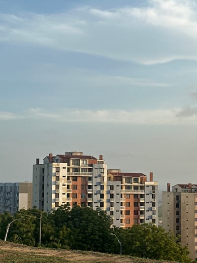 The image shows a view of apartment buildings in Perímetro Urbano Barranquilla, Colombia. The buildings are of varying heights and architectural styles, with some featuring balconies and reddish-brown brick accents. In the foreground, there are trees and a street lamp, suggesting an urban or suburban setting. The sky above is partly cloudy, with soft sunlight illuminating the scene. There are no people visible in the image.
