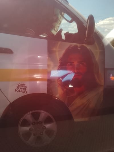 A white passenger vehicle, likely a matatu, is depicted outdoors in Kiambu Township ward, Kenya, under a clear blue sky with scattered clouds. The vehicle's side features a prominent horizontal yellow stripe. A large decal of Jesus Christ, with long brown hair, a beard, and a light-colored robe, is affixed to the vehicle's upper side, extending across a window. A faint halo or starry pattern is visible behind the depicted figure's head. A significant blue, rectangular specular reflection is present near the figure's face, possibly from a screen or reflective object. Below the yellow stripe, a smaller reddish-brown decal reads "JESUS KING KINGS" accompanied by a crown icon. A silver, multi-spoke wheel hubcap is visible at the lower left of the frame. An auxiliary rearview mirror protrudes from the upper right side of the vehicle's cab. The image exhibits a pervasive reddish-pink lens flare or reflection, particularly across the upper and central portions. No individuals or direct actions are discernible within the frame.