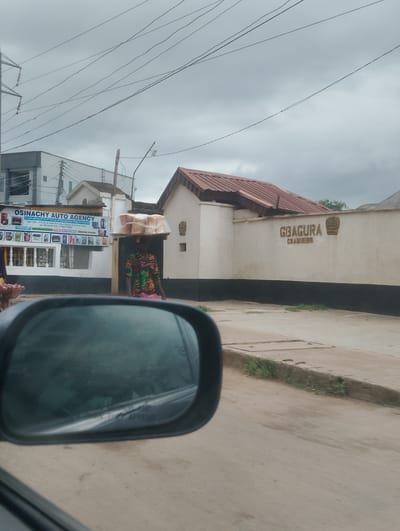 This media file captures a street scene in Iju, Nigeria. The foreground is dominated by a car's side mirror, reflecting a blurred roadway. Beyond the mirror, a woman is seen walking, balancing a large tray of bread loaves on her head. She is wearing a colorful, patterned dress.
To the left of the woman is a building with a prominent sign that reads "OSINACHY AUTO AGENCY A Subsidiary of Cecil Group of Companies Nigeria Limited". Below this text, various products like lubricants, motor batteries, tires, accessories, and general goods are advertised with small icons. The building appears to be a commercial establishment.
To the right of the woman, a white wall with black trim displays the embossed text "GBAGURA CHAMBERS". A crown-like emblem is also visible above the text. Behind this wall, a building with a reddish-brown roof can be seen.
The sky is overcast, suggesting a cloudy or possibly rainy day. The overall lighting is diffused, typical of an overcast sky, and no distinct shadows are visible, hinting at it being sometime during daylight hours, possibly late morning or afternoon. The scene depicts everyday life on a Nigerian street, with a vendor engaged in her trade and commercial buildings lining the road. The presence of power lines overhead also indicates an urban or semi-urban setting.
---
Note: The OCR output for "SO RE" is unclear and might be part of a sign or advertisement on the building to the left.