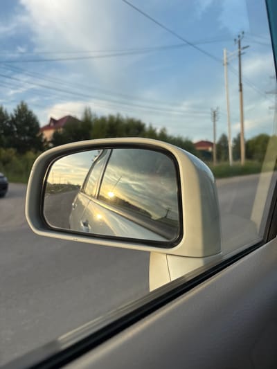 The image captures a view from inside a vehicle looking towards the side mirror. The side mirror reflects a golden sunset with clouds, and the side of the car. The exterior environment shows a road with trees and houses in the background. There are also utility poles with power lines visible. The overall scene suggests a drive through a semi-urban or rural area, possibly during the evening. The location of Chigiri, Russia is not determinable from the image itself.