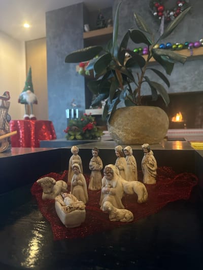 This indoor photograph depicts a Christmas Nativity scene and surrounding festive decorations within a residential space in Bend, United States.
In the immediate foreground, an off-white, multi-piece ceramic or resin Nativity set is arranged on a red, glittery mesh fabric inside a dark, rectangular container. The set includes a recumbent Baby Jesus in a manger, Mary kneeling with hands clasped, Joseph standing, a shepherd figure holding a lamb, another shepherd figure with a lamb at its feet, and five additional standing figures, totaling ten distinct pieces.
The midground features a prominent Ficus houseplant in a rounded, light brown, stone-like pot, positioned on a surface in front of a grey, textured wall.
The background reveals a decorated living area. To the left, a tall Christmas gnome figurine with a green hat, white beard, and red striped legs stands on a round table covered with a sparkly red sequined tablecloth. A partial view of a carved bird decoration is visible to its left. To the right, a lit fireplace with visible flames is embedded in the grey wall. Above the fireplace, a wooden mantelpiece displays several Christmas ornaments, including a small Christmas tree figure and a line of multi-colored spherical decorations. Below the mantel, a decorative arrangement of red flowers and green foliage, along with three white cylindrical candle holders, rests on a lower ledge.