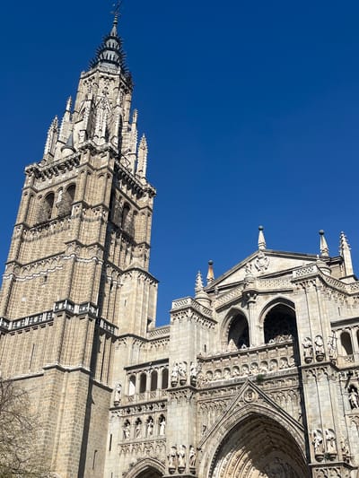 The image displays a low-angle, detailed view of the Cathedral of Toledo in Toledo, Spain, under a clear, deep blue sky. The scene is dominated by two primary architectural elements of the Gothic cathedral: its tall, multi-tiered bell tower on the left and a section of its intricately decorated facade on the right.
The bell tower, constructed from light-colored, weathered stone, rises vertically, featuring numerous architectural details including arched windows, flying buttresses, and pinnacles across its multiple levels. The tower culminates in an ornate, dark-colored spire topped with a metallic weather vane.
To the right, a section of the main facade exhibits extensive Gothic ornamentation. It features multiple pointed arches, indicative of grand portals, enveloped by elaborate relief carvings and a dense arrangement of individual statues positioned within dedicated niches along horizontal bands and vertical pilasters. Balustrades and decorative cornices further enhance the facade's complexity.
The bright daylight casts distinct shadows, highlighting the architectural depth and textures of the stone. In the lower-left foreground, several bare tree branches are visible, suggesting the presence of vegetation adjacent to the cathedral. There are no people, vehicles, or apparent actions depicted within the frame.