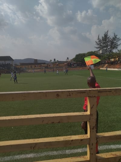 The image was taken at a soccer field in New Karu, Nigeria (latitude: 9.027, longitude: 7.578). A referee in a red shirt stands at the sidelines, raising a green and orange flag. The game is in progress. Several players are visible on the field, wearing blue and white uniforms. The spectators are sitting in the stands behind a wooden fence. The sky is partly cloudy. The field is surrounded by a fence. The image shows a match between two soccer teams. The referee is signalling something, possibly an off-side or a foul. The players are in mid-game, running and kicking the ball. The spectators are cheering and watching the game.