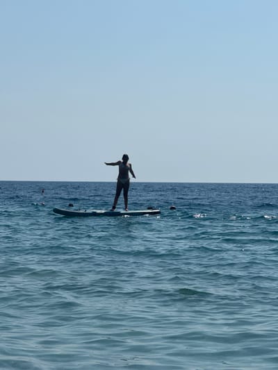 In Mirište, Montenegro, a person stands on a paddleboard in the calm, blue sea. The person, silhouetted against a clear sky, is wearing a patterned bikini. Their arms are outstretched to the sides, as if balancing. The paddleboard, which is dark in color, floats on the gently rippling water. In the distance, the horizon is a sharp line between the sea and the sky. Several buoys are visible in the water, marking a boundary or area. The overall scene depicts a peaceful moment of recreation on the water.