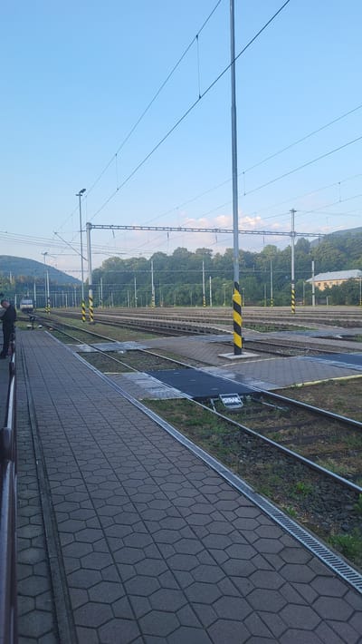 The image depicts an outdoor scene at a train station in Kysak, Slovakia. In the foreground, a paved walkway runs along the left side, with a dark railing bordering it. Beyond the walkway are multiple sets of train tracks, with gravel and grass between them. Several yellow and black striped poles are visible, likely marking hazards or points of interest. Overhead, a network of electrical wires for the trains stretches across the frame, supported by tall poles. In the middle ground, a train is visible on one of the tracks, with a person standing near the edge of the platform to the left of the image. In the background, a line of trees forms a backdrop, with a building visible further in the distance and hills beyond that. The sky is clear and blue, suggesting daylight.