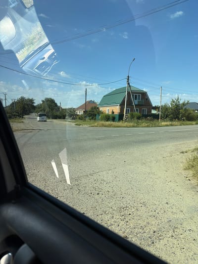 The image depicts a rural road intersection in Timashevsk, Russia, viewed from inside a vehicle. On the right side of the intersection stands a two-story brick house with a prominent green pitched roof. A lamppost is situated near the house. The road is paved with asphalt and has white lane markings. Several trees and bushes line the sides of the road, and power lines are visible overhead. In the distance, on the left side of the road, a white minivan is driving away from the viewer. The sky is blue with scattered clouds, indicating a clear day. The reflection of the vehicle's interior, including the dashboard and windshield, is visible in the foreground.