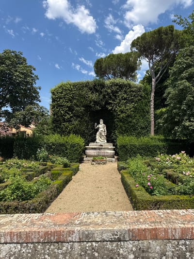 This media file captures a tranquil garden scene, likely in Florence, Italy, under a bright, sunny sky with scattered white clouds. The image is oriented vertically. The foreground features a weathered brick wall bordering a path made of fine gravel. Lush green hedges, meticulously trimmed into geometric shapes, line the path, creating a sense of structured beauty.
Deeper within the garden, a stone statue stands on a tiered pedestal. The statue appears to be of a classical figure, though its details are softened by distance and the quality of the stone. Behind the statue, a large, dense hedge forms a verdant backdrop. The garden is adorned with flowering plants, including delicate pink roses, adding pops of color to the dominant greens. The overall atmosphere is one of serene elegance and timeless charm, typical of Italian Renaissance gardens. The sunlight casts distinct shadows, suggesting it is late morning or early afternoon. No visible text is present in the image.