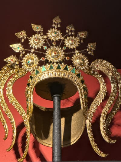 The image shows a close-up, front-facing view of an ornate golden headdress, possibly a crown, displayed on a dark stand against a dark red, textured background. The headdress is elaborately decorated with numerous small, clear gemstones and larger red and green stones. It features a central circular element with radiating points, and intricate, curved golden extensions that hang down on either side, resembling stylized wings or plumes. The headdress itself appears to be a band adorned with triangular green stones and a row of clear gemstones. The lighting casts shadows, highlighting the three-dimensional detail and metallic sheen of the object. The image is taken from a slightly low angle. There is no indication of the city of Coquitlam, Canada, in the image or its context.