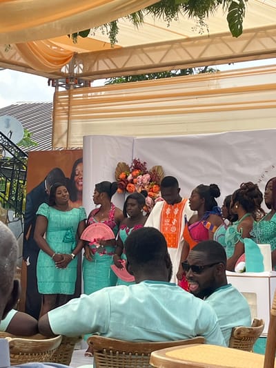 This image depicts an outdoor celebratory gathering in Kumasi, Ghana, held under a large, light-colored canopy draped with white and peach-toned fabric. In the mid-ground, a group of approximately ten individuals, predominantly women, stand against a white and peach-striped fabric backdrop adorned with a large floral arrangement composed of orange, pink, and white flowers, accented with dried palm fronds. A large portrait of a man and a woman is visible on the far left of the backdrop.
One man stands centrally within the group, wearing a white long-sleeved top with prominent orange embroidered patterns. The women wear coordinating attire, primarily consisting of light green or teal dresses and skirts, some paired with vibrantly patterned tops in shades of pink, orange, and blue. Several women hold pink, fan-shaped objects. The woman on the far left, dressed in a teal knee-length dress, stands with her hands clasped. The woman immediately to the man's right (viewer's left) wears a pink floral top and teal skirt, holding a pink fan.
In the foreground, three men are seated in wicker chairs, with their backs or sides partially visible. Two of these men wear light green/teal traditional tops, similar in hue to some of the women's garments. The man on the far right, wearing sunglasses, is turned to his left, looking towards the standing group, and appears to be speaking or smiling. Above the canopy, green foliage and a portion of a residential roof with a satellite dish are visible. A white stand with a teal decorative item or cake is present on the far right edge of the frame.