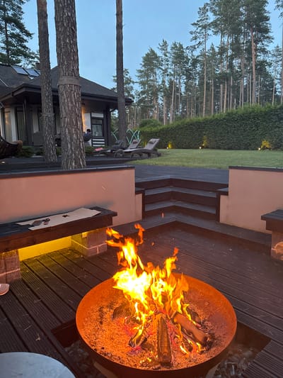 The image depicts an outdoor scene at dusk, likely in Baltezers, Latvia, as suggested by the location context. The central focus is a burning fire pit on a dark wooden deck. Flames, rendered in vibrant oranges and yellows, are actively consuming logs within the rusted metal fire pit. The deck features tiered steps leading to a grassy area bordered by a dense green hedge. In the background, tall pine trees create a natural, forest-like setting.

To the left of the deck, a modern house with dark siding and large windows is visible. Several mature pine trees are positioned close to the house, with ropes wrapped around their trunks, suggesting an attempt to preserve them or perhaps an unusual design element.

The overall mood is serene and inviting, evoking a sense of warmth and relaxation associated with an evening by the fire. The lighting indicates it's either early evening or twilight, with a clear sky. There are no visible individuals prominently featured, though subtle hints of human presence can be inferred from the setting. No text is present in the image.
