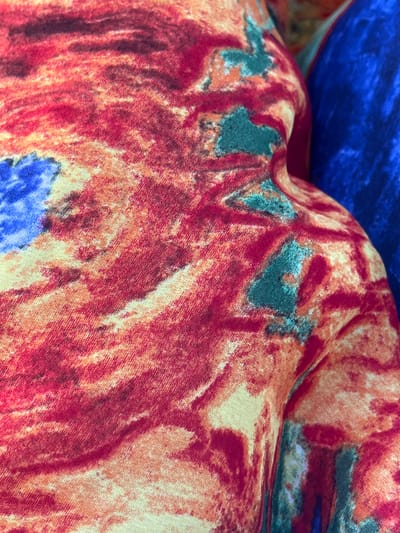 The image shows a close-up view of a textured fabric with a vibrant, abstract pattern of reds, oranges, and hints of blue and green. The fabric appears to be draped or folded, creating shadows and highlights that emphasize its texture. In the background, a section of a solid blue fabric is visible. The scene is tightly cropped, offering no context of its surroundings or location. There are no people or animals present. There is no information that can be used to determine the location in the city of Pistula, Montenegro.