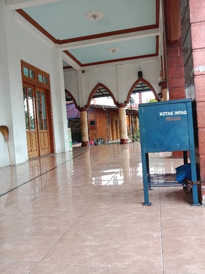 The image shows the exterior of a building in Kecamatan Brangsong, Indonesia (latitude: -6.948, longitude: 110.231). The building has a white exterior with a blue and brown ceiling. The building has a set of double doors with wood paneling. The building also has a covered portico with tiled floor. The portico is open on one side, allowing a view of a wooden building with a white roof. The tiled floor is wet and reflective, and there is a broom leaning against the wall. A blue donation box with a black metal frame is located on the right side of the image. The donation box has a sign that reads "Kotak Infaq". There is a blue cloth on the bottom shelf of the donation box. The image does not include any people.
