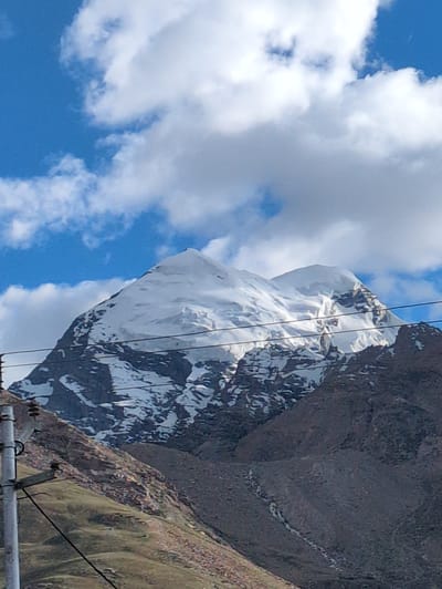 This image captures a majestic snow-capped mountain peak against a vibrant blue sky dotted with white clouds. The mountain exhibits rugged, rocky terrain with patches of snow and ice, suggesting a high-altitude environment. In the foreground, a utility pole with electrical wires is visible, indicating the presence of infrastructure near the natural landscape. The lower slopes of the mountain are covered in dry vegetation and rocky scree. The scene is likely located in or near Padum, India, a town situated in the Zanskar region of Ladakh, known for its dramatic mountainous scenery.
