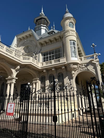 The image shows a low-angle view of the Tigre Art Museum in Tigre, Argentina. The building features ornate, light-colored architecture with towers and decorative elements. A black iron fence partially obscures the facade. A red and white sign, likely indicating a prohibition, is affixed to the fence. The sky is a clear, bright blue.