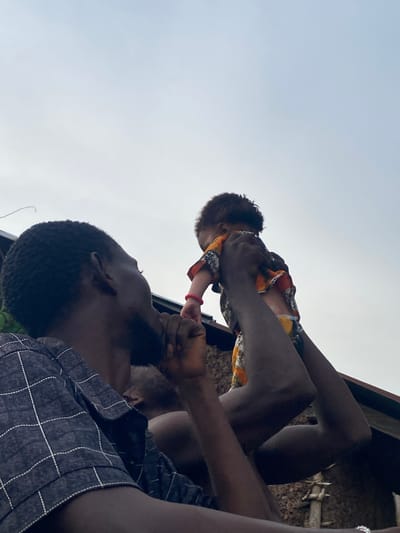 In Unwana, Nigeria, a low-angle, medium shot captures a man holding a baby aloft against a pale sky. The man, seen from the side, wears a dark, plaid shirt and rests his chin on his hand. His eyes are directed upwards towards the baby. The baby, adorned in a brightly colored, patterned garment with a red bracelet on its left wrist, is held by the man's outstretched arms, its back to the camera. The scene is set outdoors, with hints of rustic structures and possibly vegetation in the background, suggesting a simple, domestic environment. The lighting is diffused, indicating an overcast day.