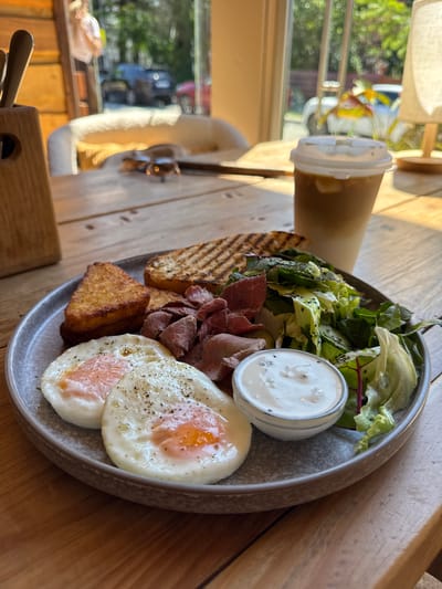 A close-up, eye-level, vertical shot showcases a breakfast plate on a rustic wooden table. The plate holds two fried eggs with visible yolks, seasoned with black pepper, a triangular hash brown, thin slices of roast beef, grilled toast, a small salad with leafy greens and sliced cucumber, and a ramekin of white sauce. To the right of the plate is a tall to-go cup with a white lid, containing a light brown beverage with ice. In the blurred background, sunlight streams through large windows, illuminating a chair with a fluffy white seat, a wooden utensil holder with cutlery, and a lamp with a beige shade. Outside the window, foliage and vehicles are visible, suggesting an outdoor or semi-outdoor dining setting. The overall scene depicts a breakfast meal in a cafe or restaurant environment, possibly in Krasnaya Polyana, Russia, given the ambient light and natural elements.