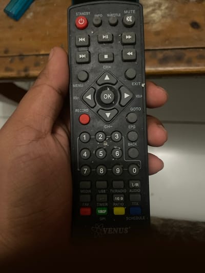 A person's right hand holds a black remote control. The remote is a Venus brand and has buttons for various functions, including standby, info, subtitle, mute, record, menu, exit, volume, channel, goto, epg, back, media, usb, tv/radio, l/r audio, fav, timer, 16:9 ratio, ttx, dpi, 1080p, and schedule. The hand is holding the remote in front of a wooden surface. The background of the photo is a white wall. The scene appears to have been captured indoors. The coordinates provided correspond to a location in Cimenyan, Indonesia. It is unknown why the person is holding the remote, but there is no evidence of any other actions or interactions in the image.