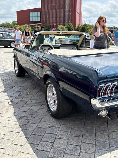 A dark blue convertible classic car, likely a Ford Mustang, is parked on a cobblestone surface. The car is the primary subject, with its rear driver's side visible. The car has chrome accents and shiny rims on its wheels. In the background, several people are present, including a woman with red hair wearing sunglasses and a grey t-shirt, standing near another classic car. There are buildings in the background, one of which is tall and brick-colored with a modern design. The environment appears to be an outdoor event or gathering, possibly a car show, taking place on a cloudy day with sunlight casting shadows. The scene is set on a paved area that resembles a city square or promenade, consistent with locations such as the city of Bruges, Belgium, known for its historic architecture and public spaces.