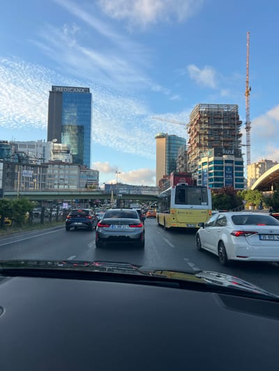 This is a vertical, slightly tilted, eye-level shot of a busy street scene in Beşiktaş, Turkey. The image captures a traffic jam with several cars and a yellow bus stopped on a multi-lane road. The architecture is modern, with several tall buildings visible, including one with "MEDICANA" prominently displayed and another under construction with a large crane. Other visible text includes "TEKYOL," "fuzul," "Sofrada," and "Coca-Cola," suggesting various businesses and advertisements. The sky is a bright blue with wispy white clouds, indicating it's daytime, likely late afternoon or early evening given the lighting. The overall atmosphere appears to be one of urban hustle and bustle, characteristic of a major city.