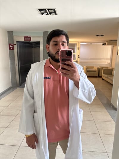 A man in a white coat and peach polo shirt is taking a mirror selfie. He holds his phone up to the mirror, partially obscuring his face. His dark hair and beard are visible. The reflection shows a hallway with an elevator and seating area in the background. Tile floors, white walls, and recessed lighting are present. The text "ruca" is visible on the polo shirt. A gold watch is seen on his wrist.
