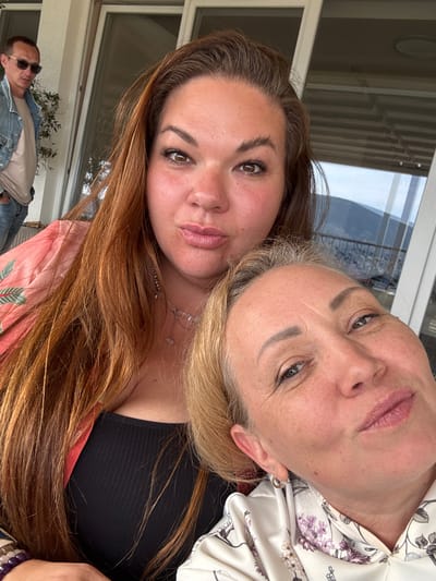 The image shows two women in a close selfie pose. The woman on the left has long, auburn-colored hair, while the woman on the right has blonde hair and is wearing a light-colored floral blouse. A third person is visible in the background, a man wearing sunglasses and a denim jacket is lying down. The location appears to be on a balcony. It is not possible to verify or deny the city of Trebesin, Montenegro based on the image alone.