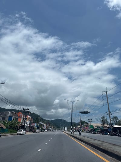 The image shows a wide, paved road with a yellow line marking the edge of the lane. The road is lined with buildings on both sides, appearing to be a commercial or residential area in Ban Khuk Khak, Thailand. There are several parked cars and motorcycles along the roadside. Power lines and streetlights are visible throughout the scene. In the background, there are hills covered in vegetation. The sky is partly cloudy with patches of blue sky visible.