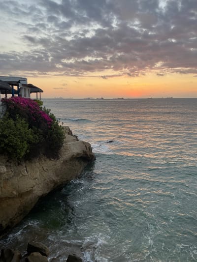 The image shows a scenic view of the ocean at sunset in Ballenita, Ecuador. A cliffside structure with a thatched roof and white walls is visible on the left, adorned with vibrant pink bougainvillea flowers and lush green foliage. The structure overlooks the ocean, where several cargo ships are dotted on the horizon. The sky displays a gradient of colors, transitioning from soft grays and pinks of the clouds at the top to a warm orange and yellow glow of the setting sun near the horizon. The ocean surface reflects the colors of the sky, with gentle waves lapping against the rocky shore at the base of the cliff. The overall atmosphere is tranquil and picturesque.