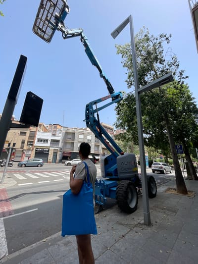 A person stands on a sidewalk, carrying a bright blue tote bag. They are facing away from the camera, looking towards a large blue aerial work platform, also known as a cherry picker. The platform's boom is extended upwards, with a basket at the end. In the background, a street is visible with cars, buildings, and trees lining the sidewalk. The scene appears to be in an urban environment.
The time of day is daytime, with bright sunlight casting shadows, suggesting a clear day. The presence of the aerial work platform indicates that some kind of maintenance or construction work is likely taking place, possibly on the streetlights or the buildings.
Visible text on the aerial work platform includes "ALTE GX 150/26" and "ALQUILER" (Spanish for "rental"). There is also a "P" sign with a parking symbol visible in the background. The location context of Santa Coloma de Gramenet, Spain, is provided.
