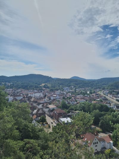This is a high-angle, full-frame photograph of the town of Dahn, Germany, taken on a bright, partly cloudy day. The foreground is dominated by lush green pine trees, partially obscuring the view of the town below. The middle ground reveals a dense cluster of red-tiled roofs belonging to houses and buildings, interspersed with trees. A prominent white church with a tall steeple is visible in the center of the town. A road with parked cars winds through the town, and a railway line can be seen on the right side of the frame. The background features rolling, tree-covered hills and a hazy blue sky with scattered white clouds. The overall impression is a serene, pastoral view of a German town nestled within a natural landscape.