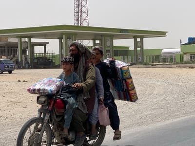 A red motorcycle transports four males, one adult and three children, traveling from right to left on a paved road in Daman, Afghanistan. The adult male, with a beard and wearing a turban, operates the motorcycle. A young boy in a patterned skullcap is positioned in front of the driver, gripping the handlebars, with a clear plastic bag containing numerous small packages affixed to the front of the motorcycle. A second child is partially obscured between the driver and the rear passenger. The rear passenger, a boy wearing dark clothing and sandals, carries a large, plastic-wrapped stack of snack bags over his left shoulder and holds a pink plastic bag in his left hand.
The background features a green and white gas station. This station includes a multi-pillar canopy structure, fuel pumps, and a white horizontal LPG storage tank. An additional green and white building is visible to the right of the LPG tank. Behind the gas station, a tall, reddish-brown metal lattice communication tower extends into the hazy, light blue sky. To the left of the gas station, a light blue van with roof-mounted lights is present on the unpaved, dusty ground.
