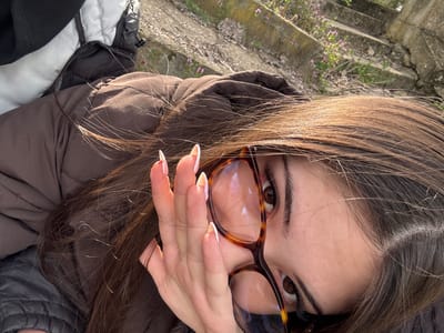 A young woman with long brown hair and brown eyes is angled in the frame. She wears glasses with tortoise-shell frames. Her hand is raised near her face, displaying long, pale fingernails with white tips. She wears a brown jacket. The background shows a weathered stone structure with green grass and small purple flowers, suggesting a potentially historical or old outdoor setting. The image is taken from a tilted perspective. There is no indication of the specific location in Gromshin, Bulgaria.