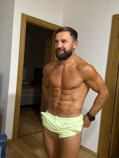 A man with a beard and tan skin stands in front of an open doorway. He has defined abdominal muscles and is wearing light yellow shorts. He is looking to his left. A watch is visible on his left wrist. The room behind him has a bed and wooden flooring. The wall to his left is white. The scene appears to be indoors.