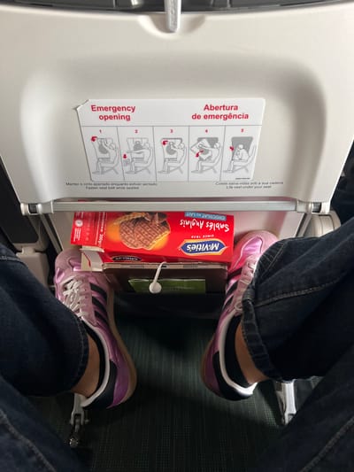 The image shows the leg and shoe of a person on an airplane. The person is wearing dark blue jeans and pink and black athletic shoes. In front of the person's legs is a tray table, on which a box of McVitie's chocolate biscuits and a tablet are placed. An emergency instruction card is visible on the back of the seat in front of the person. The location is the cabin of an airplane, in Paray-Vieille-Poste, France.