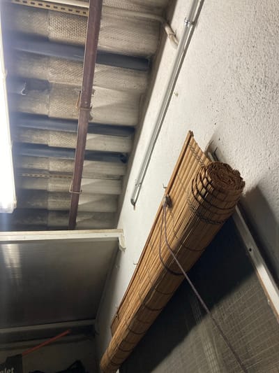 The image shows a close-up view of an interior space, likely a garage or storage area, in Sabadell, Spain. The scene is dominated by a rolled-up bamboo blind mounted on a textured white wall. Above the blind, a corrugated ceiling made of light-colored material is visible, supported by dark horizontal beams and a rusty metal pipe running vertically. To the left, a fluorescent light fixture is partially visible. A metal frame structure, possibly a shelf or partition, extends across the bottom left of the frame. The overall impression is one of a utilitarian, perhaps unfinished, space.