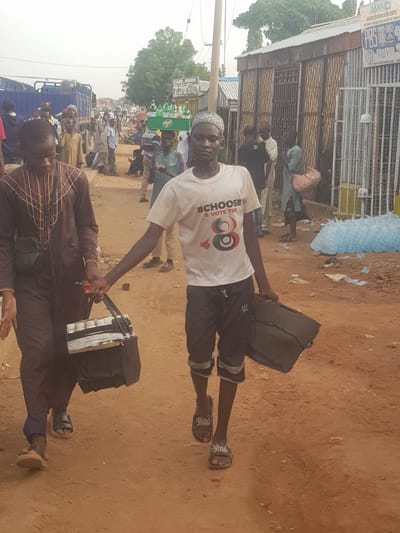 The scene is an outdoor setting, possibly a street or market area in Kufena, Nigeria. The ground is mostly bare earth with a reddish hue. People are scattered throughout the frame, suggesting a public gathering or market. A young man wearing a white t-shirt with political messaging ("#CHOOSE & VOTE THE 8") is holding two black rectangular objects, possibly bags or cases. He is also wearing shorts and sandals. Another man in a brown patterned outfit is in the foreground holding one of the black bags. There is a building with a fenced-in storefront on the left side of the image, and a blue truck in the middle of a crowd. There are trees and general urban clutter scattered throughout the scene.