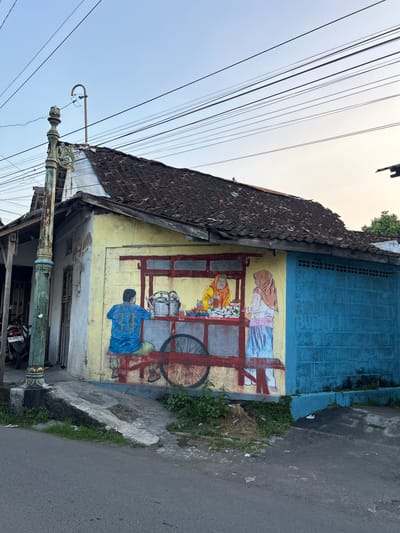 The image captures a vibrant mural painted on the side of a yellow building, depicting a street scene. The mural features two women and a child, seemingly engaged in a transaction. The woman on the right, wearing a light pink hijab and a blue and white outfit, is bent over a cart laden with goods. A child in a bright blue shirt with yellow numbers is also present, kneeling and reaching towards the cart. The cart itself is a prominent element, painted in red and featuring a large black wheel. Behind the mural, the building's facade is painted in a contrasting bright blue with a brick pattern. The roof is tiled with dark brown tiles, and a section of the building's wall shows signs of wear and tear.
The setting appears to be a street in Yogyakarta, Indonesia, suggested by the presence of overhead power lines, a traditional-looking street lamp post with ornate details, and a motorcycle parked near the building's entrance. The road in front of the building is asphalt. Green vegetation grows along the edge of the sidewalk and against the building.
The mural's bright colors and the depiction of everyday life evoke a sense of community and economic activity. The time of day appears to be late afternoon or early evening, given the soft, warm light and the long shadows. The sky is a pale blue, suggesting clear weather. There is a license plate visible on the motorcycle with the text "AB 6860 GL."