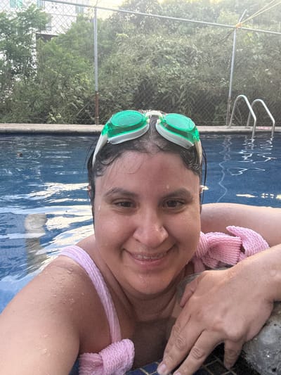A person is submerged up to their shoulders in a swimming pool. They are wearing swimming goggles. The pool appears to be outdoor, bordered by foliage and a chain-link fence. A ladder for pool access is visible at the edge of the pool. The location may be Cuernavaca, Mexico, although this is difficult to confirm from the image alone.