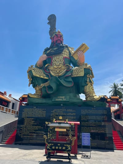 The image shows a large, imposing statue of Guan Yu, a historical Chinese military figure, seated and holding a book. The statue is painted in green and gold, with a red face and a long black beard. It is erected on a black monument with inscriptions in Thai and Chinese. In front of the monument, there is an ornate incense burner, also with Chinese inscriptions. The background features a clear blue sky with some scattered clouds, palm trees, and the facade of a traditional Chinese-style building with red lanterns. The location is identified as the city of Baan Hua Thanon, Thailand, with the statue being a prominent landmark. The scene appears to be an outdoor setting on a sunny day, possibly a temple or a cultural site. There are no people directly visible in the frame interacting with the statue or the monument.