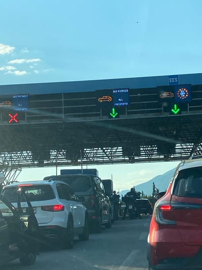The image shows a line of cars at a border crossing point, likely in Palje Brdo, Croatia, as indicated by the context. The traffic is heavy. Overhead signs indicate lane designation. The upper sign displays the EU flag and "EES" with a car symbol and a green arrow. Another sign shows "SVI PUTNICI" (All Passengers) and "ALL PASSPORTS" with a car symbol and a green arrow. The final sign reads "SVI PUTNICI" and has "ALL PASSPORTS" along with a car symbol and a red X. The weather appears clear and sunny. The scene suggests a potential delay for travelers crossing the border due to the volume of traffic and varying passport requirements based on the signage.