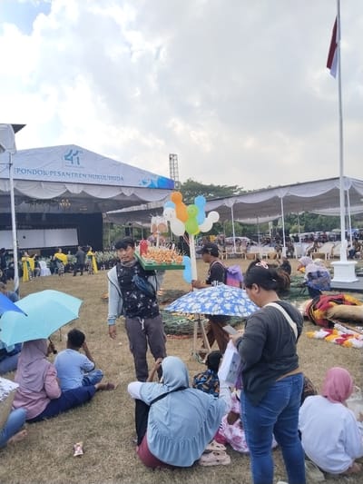 In Plosorejo, Indonesia, an outdoor event unfolds under a cloudy sky, likely a celebration or festival at Pondok Pesantren Nurul Huda, indicated by banners. A vendor in the foreground carries a tray of food items, possibly snacks, and is accompanied by several colorful balloons. In the mid-ground, a stage is set up with performers in traditional attire, suggesting a cultural or religious presentation. A diverse crowd is seated on the grass, some under umbrellas, observing the activities. The overall atmosphere appears relaxed and communal, with attendees enjoying the festivities in the open-air setting.