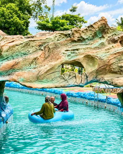 In the city of Eampat Kwalamencirim, Indonesia, three individuals are enjoying a lazy river ride at a water park. Two women and one man are seated in a blue inflatable tube as it floats through clear, turquoise water. The woman on the left wears a green headscarf and shirt, while the woman on the right is dressed in a burgundy headscarf and shirt. The man, in the center, is smiling. The lazy river is bordered by a blue tiled wall and a large, artificial rock formation that resembles a cave entrance, painted in earthy tones with streaks of green and blue. Lush green trees and a partly cloudy sky are visible in the background.