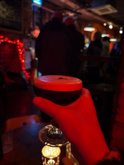 A hand holding a glass filled with a dark liquid is viewed from an unusual angle. The hand is bathed in red light, as is the rim of the glass. The glass base is visible, reflecting the light from nearby lamps. In the background, a series of blurred red lights are strung across a dark, partially obscured scene. This suggests a festive or decorative setting, though the details are indistinct.