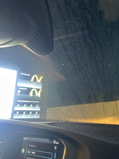The image depicts the interior of a stationary vehicle looking outward through a heavily soiled windshield during the evening. The lower portion of the frame shows the vehicle's dark dashboard, with a prominent infotainment screen in the lower-left corner displaying "20:17" (time), "0 km/h" (speed), and "89.7 RFM" (a radio frequency, consistent with Portuguese broadcasting). In the upper-center, a black plastic component, possibly an air vent or sensor housing, is visible.
Beyond the windshield, to the left, a brightly illuminated digital McDonald's drive-thru menu board is visible, displaying the iconic golden "M" arches twice, vertically stacked. Below these, rows of beverage icons (cups with straws, coffee cups) are presented. The exterior on the right is dark and indistinct, with horizontal light-colored banding at its base.
The scene takes place at a McDonald's drive-thru located at Rua da Cruz d'Areia 180, 4800-410 Guimarães, Portugal. No individuals are visible. The vehicle is static, facing the menu board, indicating an ordering process is underway.