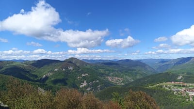 A scenic view from Sini Vrah, Bulgaria reveals a lush mountain range under a bright blue sky dotted with fluffy white clouds. The foreground features a cluster of trees with reddish-brown leaves. The mountains are covered in green forests, with hints of rocky outcroppings and a small village nestled within one of the valleys. To the right, a settlement with buildings and a large structure, possibly a monastery, can be seen. The landscape stretches into the distance, creating a sense of depth and vastness.