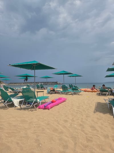 The image depicts a beach scene in Dyuni, Bulgaria, under a cloudy sky. The foreground is dominated by sand and several rows of empty green beach chairs with teal umbrellas. In the middle ground, two children, a girl and a boy, are sitting on an inflatable raft. To their right, a man is partially visible on a lounge chair. Further back, other beach chairs and umbrellas are scattered across the sand. The sea is visible in the background, with a breakwater and a lighthouse in the distance. Several people are in the water. The overall atmosphere suggests a cloudy day at the beach.