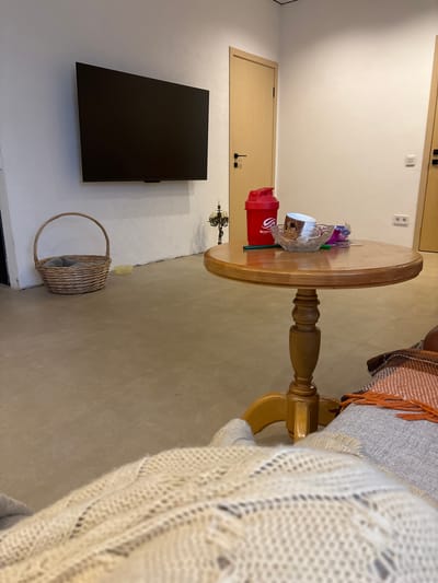 The image depicts an indoor scene, likely a living room, with a warm, neutral color palette. A large, flat-screen television is mounted on a textured white wall. To the left, a woven basket sits on the floor. In the center of the room, a round wooden table with a pedestal base holds a red shaker bottle, a glass bowl containing some items, and what appears to be paper or plastic wrappers. A candlestick holder with multiple candle sockets stands to the left of the table. Two wooden doors are visible in the background, one slightly ajar. The flooring is tiled in a light brown or beige color. In the foreground, a person is partially visible, covered by a textured white blanket and a patterned throw blanket. The setting does not provide specific clues to its location within Vardane-Verino, Russia.