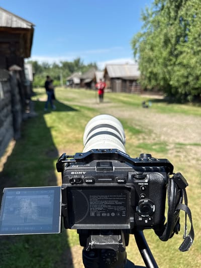 The image shows a Sony FX3 Cinema Line camera with a telephoto lens mounted on a tripod. The camera is aimed at a village scene with traditional wooden houses and a grassy path. In the background, two people are walking away from the camera, and another person in a red shirt is visible further down the path. The environment appears to be outdoors on a sunny day. The location is identified as Shushenskoye, Russia. The camera's LCD screen shows a preview of the scene.