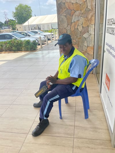 The image depicts a security guard seated on a blue plastic chair outdoors, possibly in the city of Unguwar Rimi, Nigeria. The guard, wearing a dark baseball cap, a light blue shirt, a neon yellow safety vest, and dark blue trousers with a light blue stripe, holds a black and yellow Garrett metal detector in their lap. The guard's left hand rests on the metal detector. The background shows a wall constructed of irregularly shaped stones of various earth tones, and to the right, a white vertical sign with text and a red rectangle. In the distance, several cars are parked in front of a large white tent. The ground is paved with beige tiles. The lighting suggests it is daytime with some cloud cover.