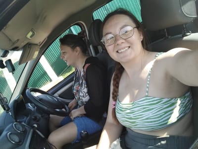 Two women are in the front seats of a car, likely in Блохино, Russia. The woman in the passenger seat is looking out the window to the left, while the woman in the driver's seat is looking at the camera and smiling. She is wearing glasses and a green and white striped bikini top. Her hair is in a braid. The woman in the passenger seat has her hair in a bun and is wearing a black jacket over a colorful shirt. The car's interior is visible, including the steering wheel, dashboard, and a gear shift. The weather appears to be sunny, with bright light streaming into the car. The scene is set during the day.